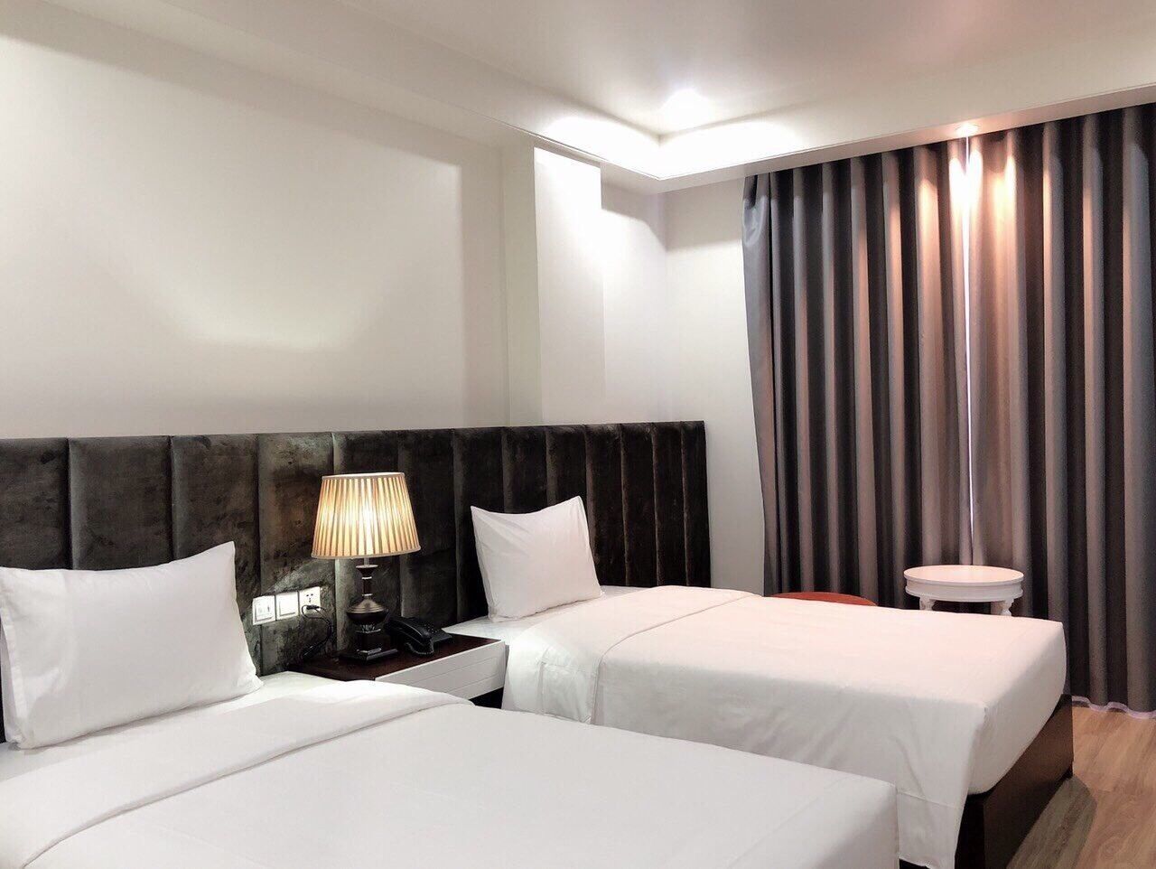 Habitación de hotel con dos camas, una mesita de noche con lámpara, una pequeña mesa y un amplio ventanal con cortinas.