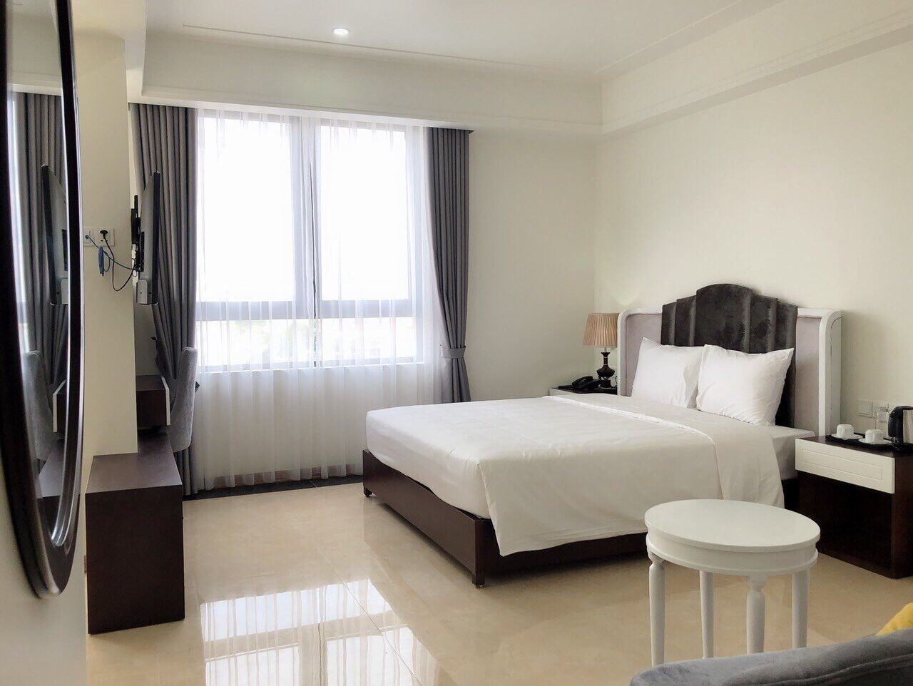 A hotel room with a large bed, a bedside table, a nightstand, a mirror, and a window with curtains.