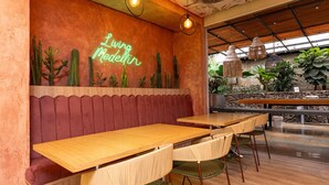 Interior de restaurante con mesas de madera, sillas tejidas, un letrero de neón y un mural de cactus.