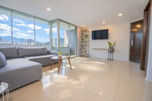 Een moderne woonkamer met een grijze hoekbank, een klein rond tafeltje, een potplant, een aan de muur bevestigde televisie en een schuifdeur die leidt naar een balkon.