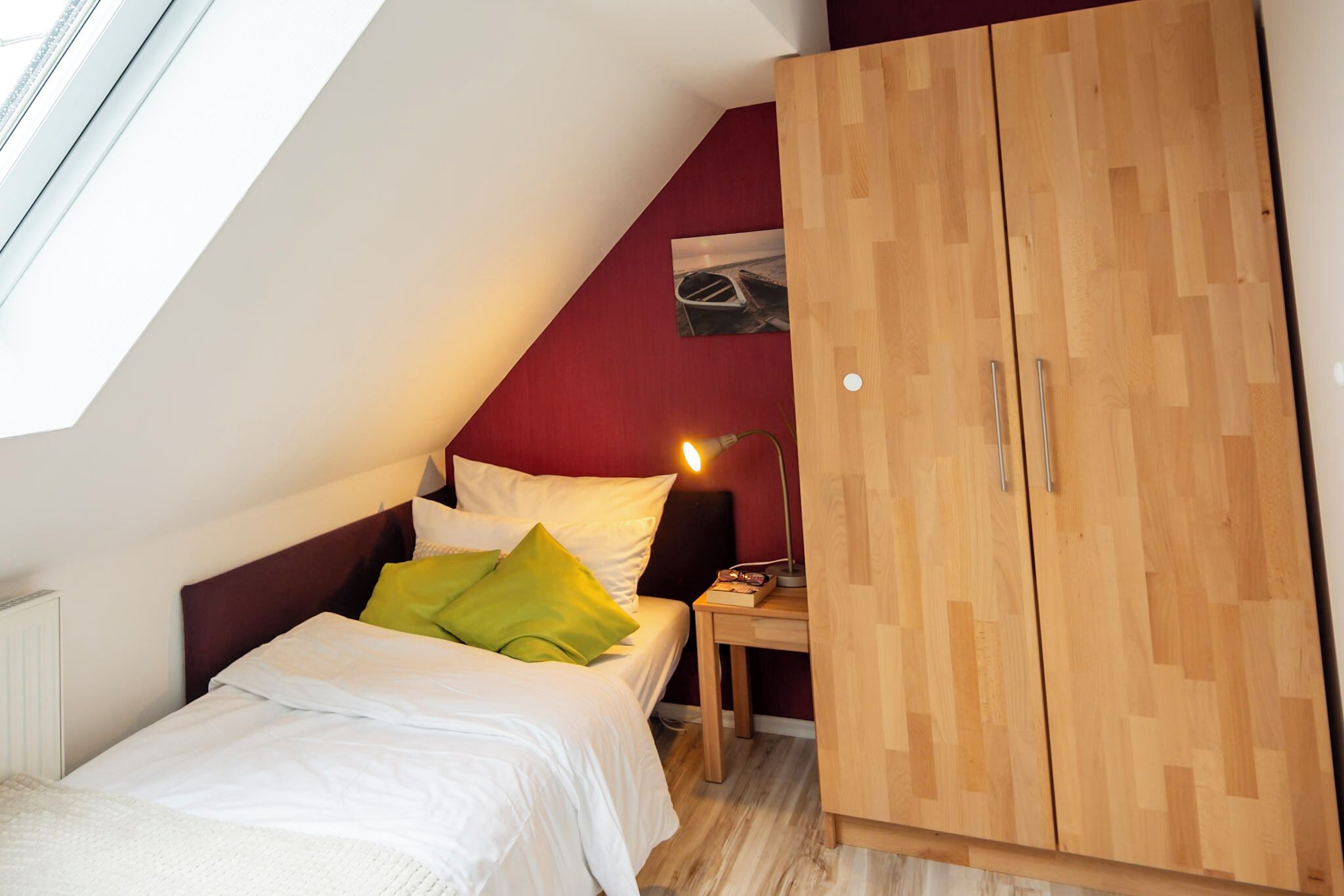 Un dormitorio con techo inclinado, cama con ropa de cama blanca y almohadas verdes, mesita de noche con lámpara y un armario de madera.