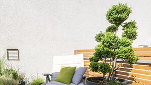 En seng med hvid madras og puder, en stor plantepotte og et træhegn i en patio.