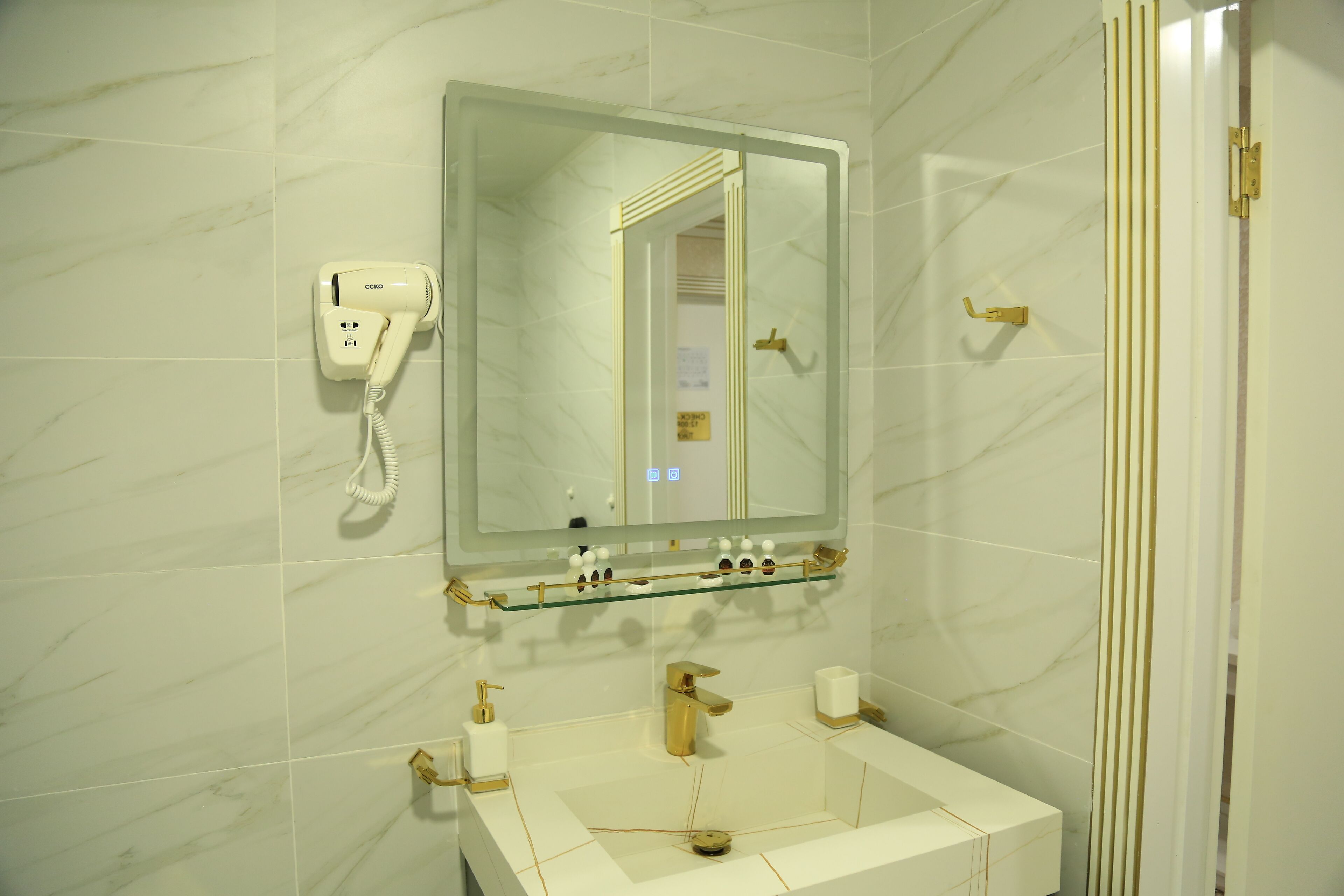 A bathroom with a marble wall, a glass shelf with toiletries, a gold faucet, and a hairdryer on the wall.