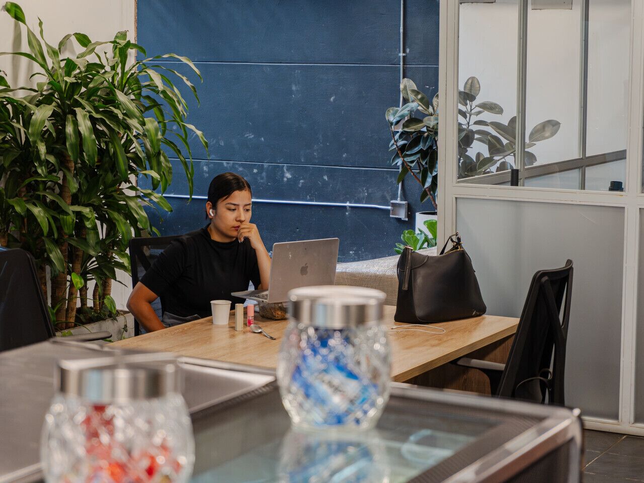A person working at a wooden table with a laptop, surrounded by plants and decorative items.