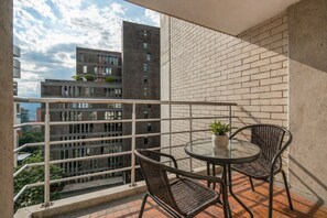 A balcony with a glass table and wicker chairs, a potted plant, and a view of a modern building.