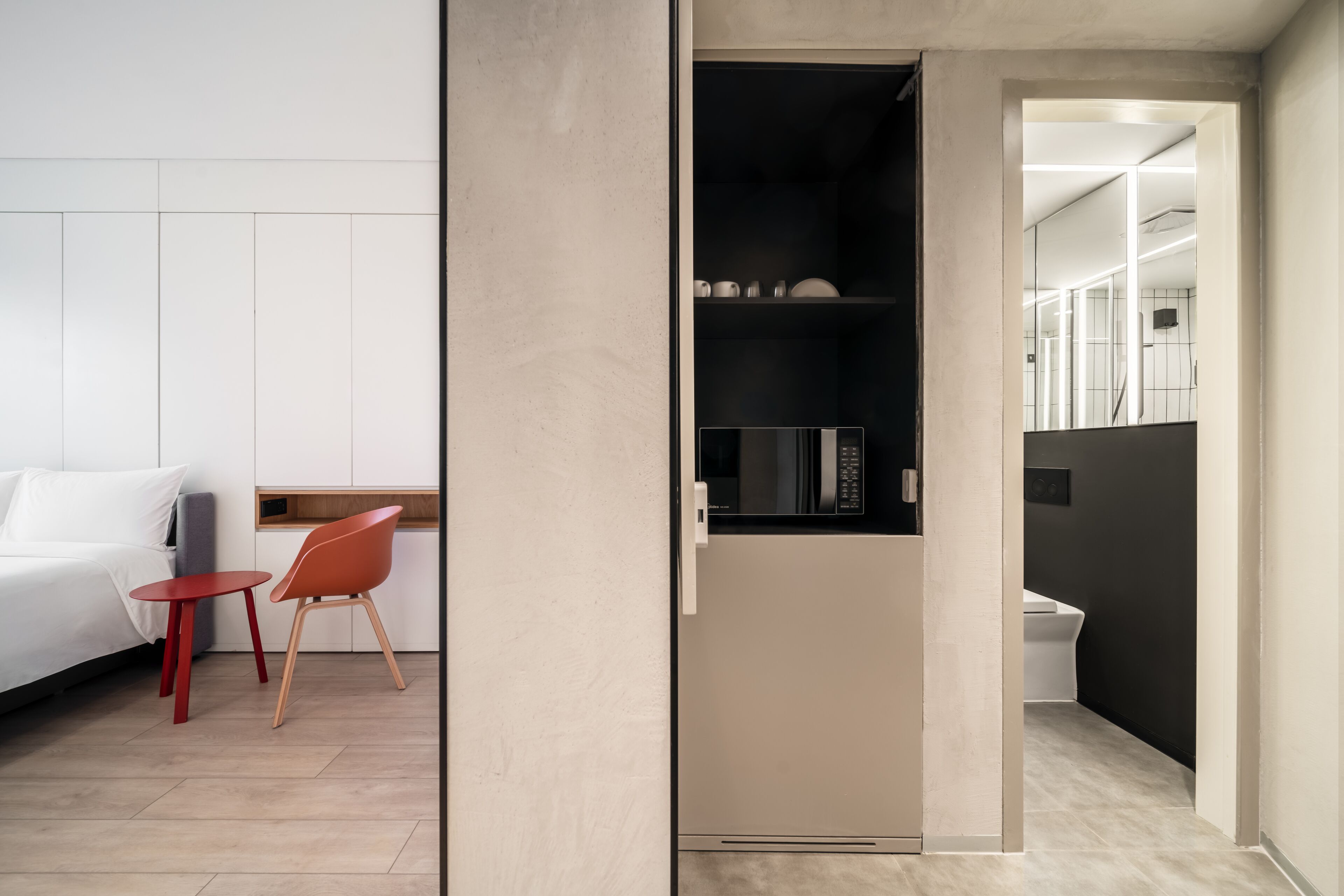Een moderne hotelkamer met een bed, een klein rode tafel, een rode stoel en een kitchenette met een magnetron.