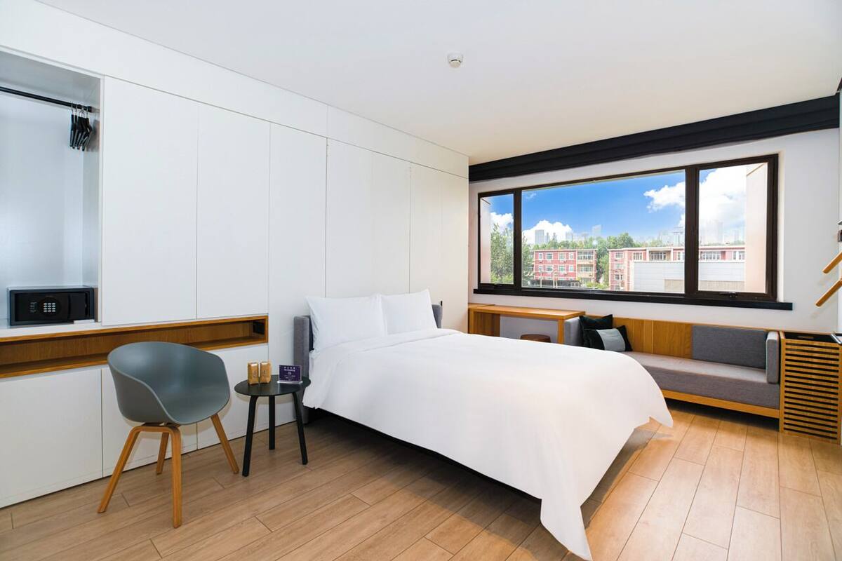 Une chambre d’hôtel moderne comprenant un lit, un coin salon avec des coussins, une petite table avec une chaise, et une vue sur les bâtiments à travers de grandes fenêtres.