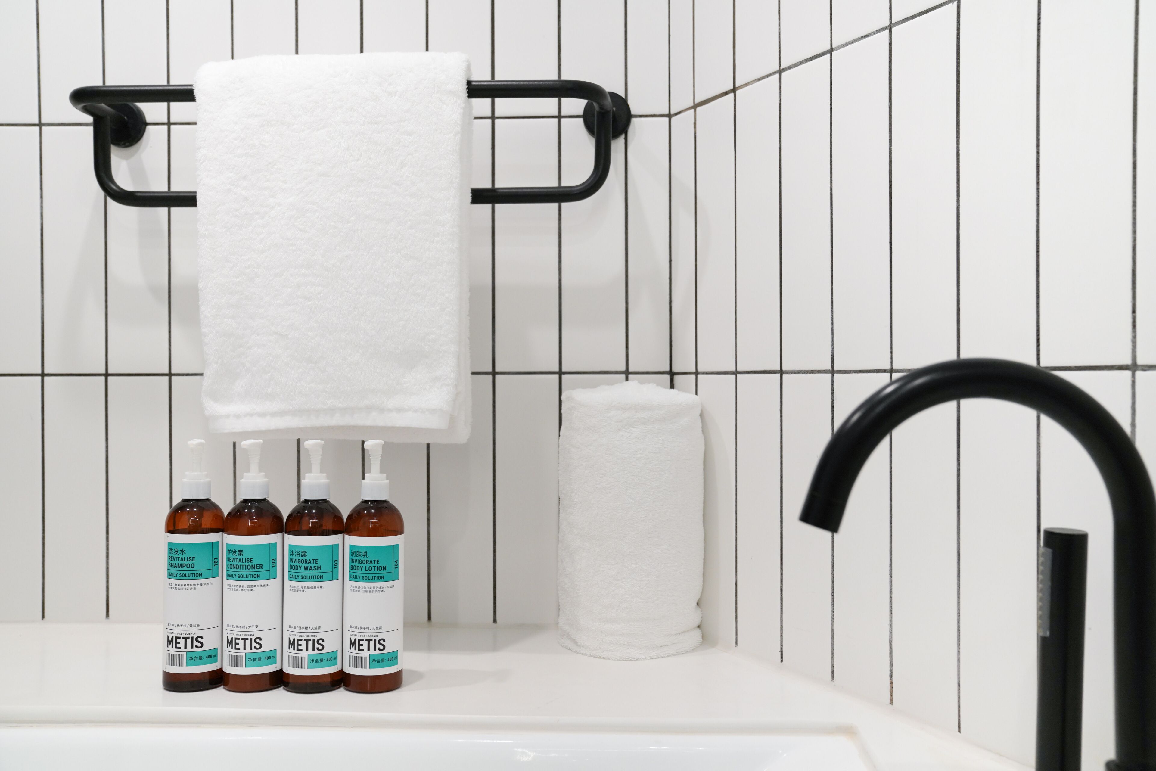 A bathroom with a white sink, four bottles of METIS body wash, a black towel rack, and a black faucet.