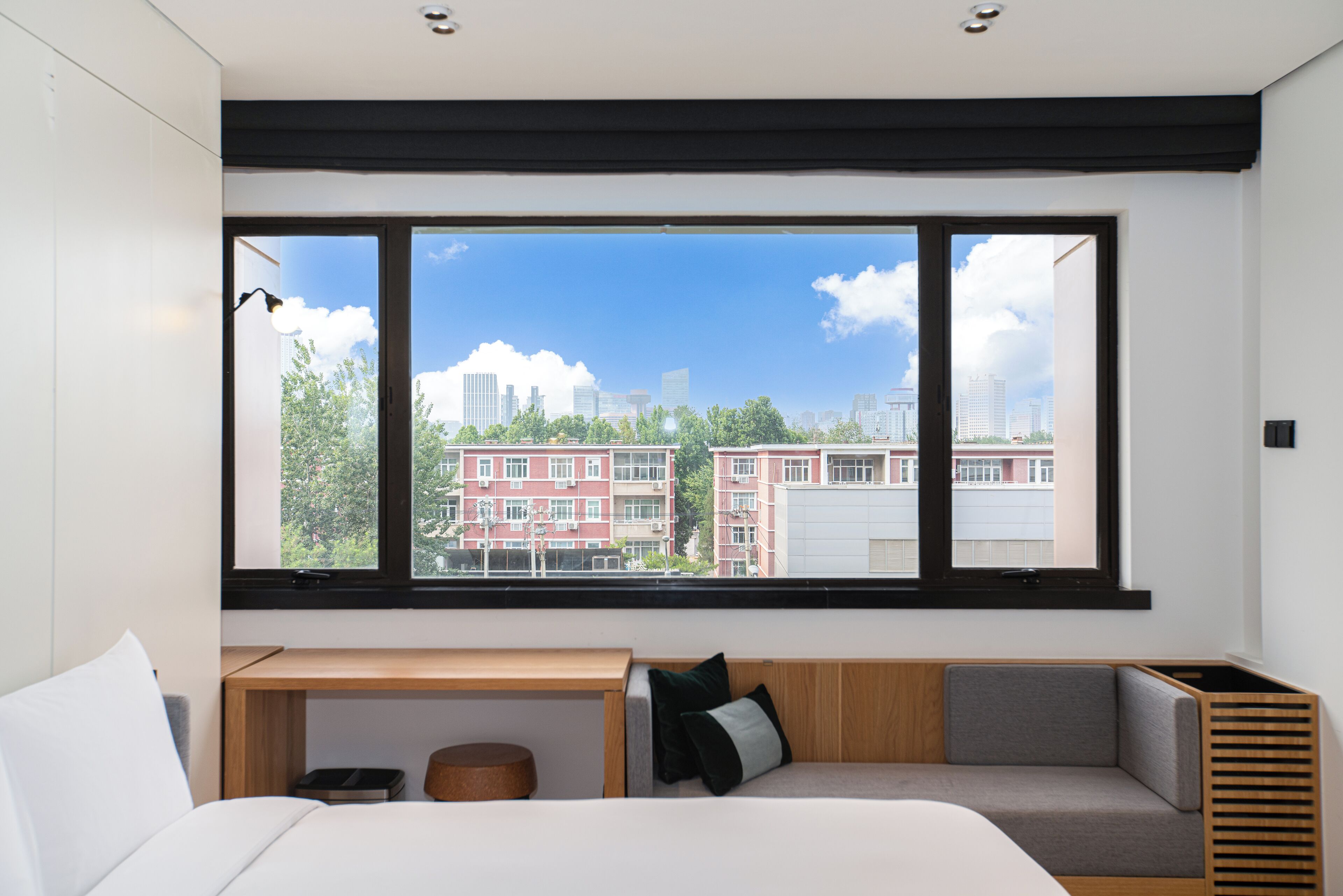 A modern hotel room with a large window, a bed, a wooden desk, and a seating area.