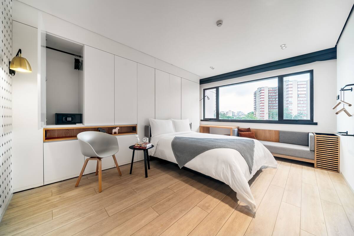Une chambre moderne avec un lit, des tables de chevet, une chaise, une fenêtre et une vue sur des immeubles.