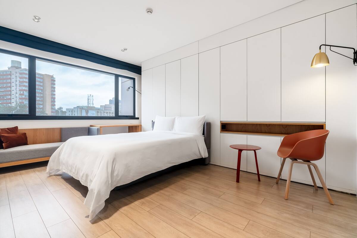 Une chambre d’hôtel moderne avec un grand lit, un coin salon comprenant un banc, une petite table et une chaise. Une fenêtre offre une vue sur les bâtiments environnants.
