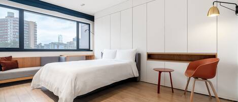 Habitación de hotel moderna con una cama grande, una zona de estar con un banco, una mesita pequeña y una silla. Se aprecia una ventana con vistas a edificios.