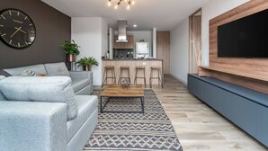 A modern living room with a grey sofa, a coffee table, and a patterned rug. There is a kitchen area with bar stools and a large TV mounted on the wall. - Go Living 708 · Luxury Apt-balcony and city view-AC and coworking (Medellín)