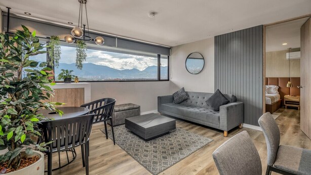 Un salon moderne avec un canapé gris, une table basse ronde et un coin repas comprenant une table et des chaises. On y trouve également des plantes, une grande fenêtre donnant sur les montagnes et un miroir accroché au mur.