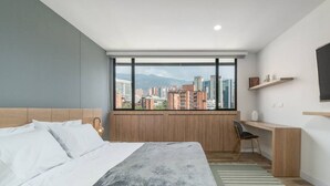 A modern bedroom with a large bed, a desk, and a view of a cityscape through a large window. - Go Living 908 · Modern Loft in El Poblado w/ AC, GYM and Cowork (Medellín)