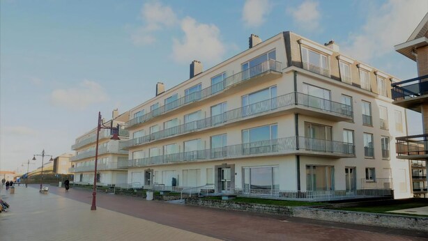 Un edificio residencial de varios pisos con balcones, un paseo pavimentado y cielo despejado.