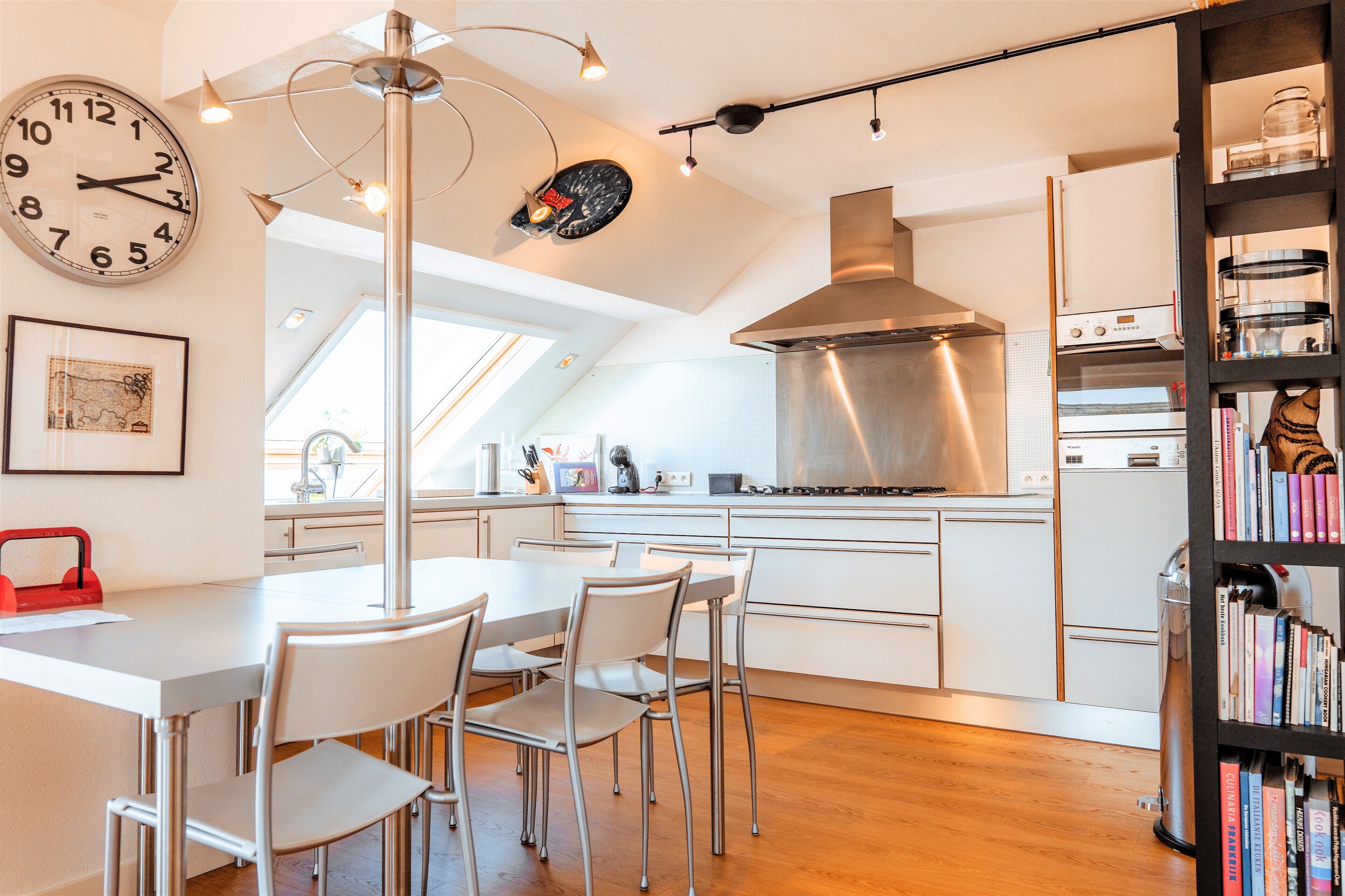 A modern kitchen with a dining area, featuring a clock, pendant lights, and a bookshelf.