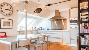 Una cocina moderna con comedor, que incluye un reloj, lámparas colgantes y una estantería.