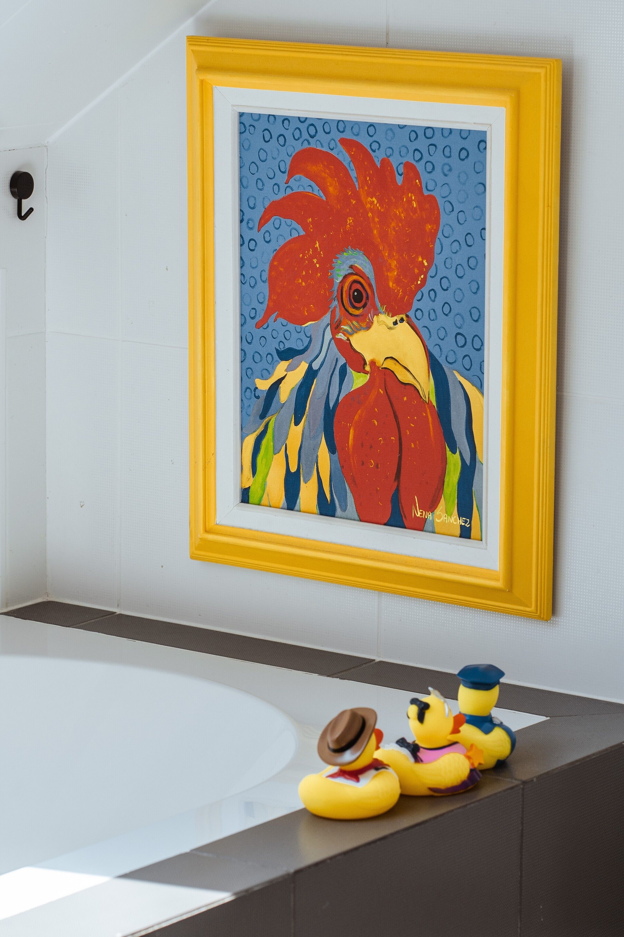 A framed painting of a rooster on a wall, a bathtub, and rubber ducks with hats.