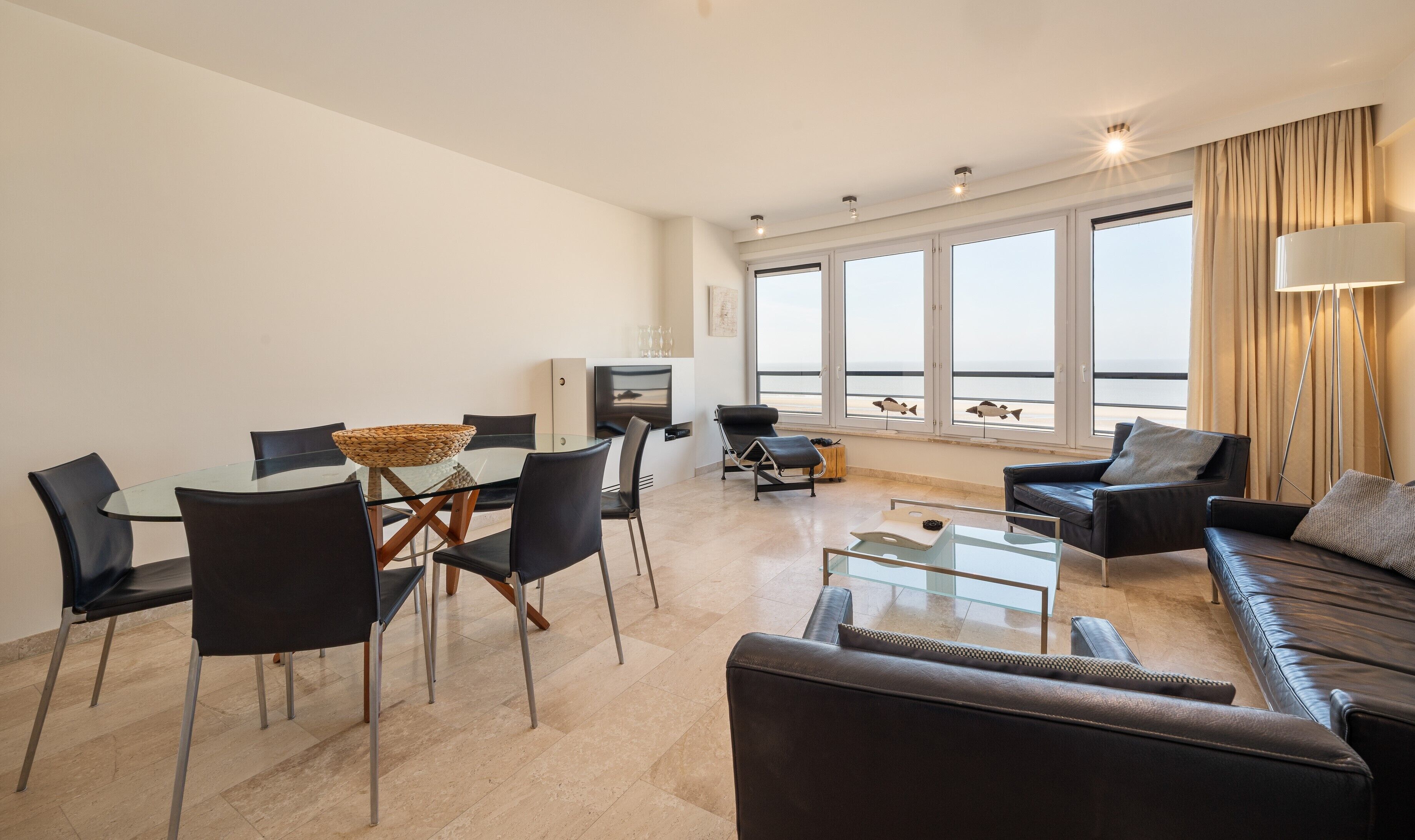 Een moderne woonkamer met een glazen eettafel, zwarte leren banken en een groot raam met uitzicht op zee.