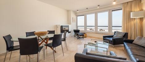 Een moderne woonkamer met een glazen eettafel, zwarte leren banken en een groot raam met uitzicht op zee.