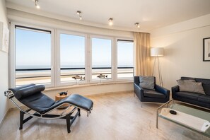 A modern living room with a large window overlooking the sea, a leather recliner, a dark sofa, and a glass coffee table. - Cockpit 0301 - beautiful 2-bedroom apartment with sea view (De Haan)