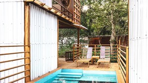 Zona de piscina con terraza de madera y sillas, rodeada por una valla metálica y árboles.