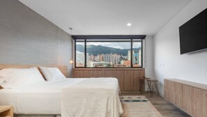 A modern hotel room with a large bed, a small table, a TV, and a city view through a large window.