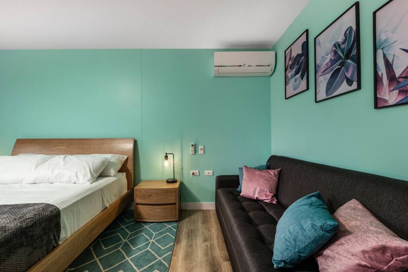 A room with a bed, a bedside table with a lamp, a couch with pillows, an air conditioner, and wall art.