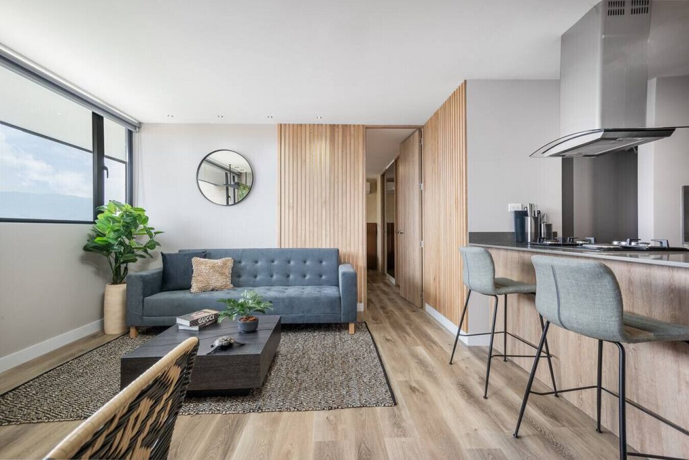 A modern living room with a grey sofa, a coffee table, and a small plant. There is a kitchen area with a bar counter and stools.