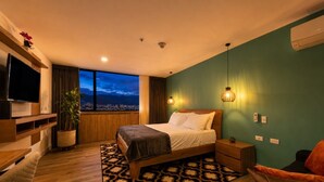 Moderni hotellihuone, jossa on suuri sänky, televisio, ruukkukasvi ja näkymä kaupungin siluettiin auringonlaskun aikaan.