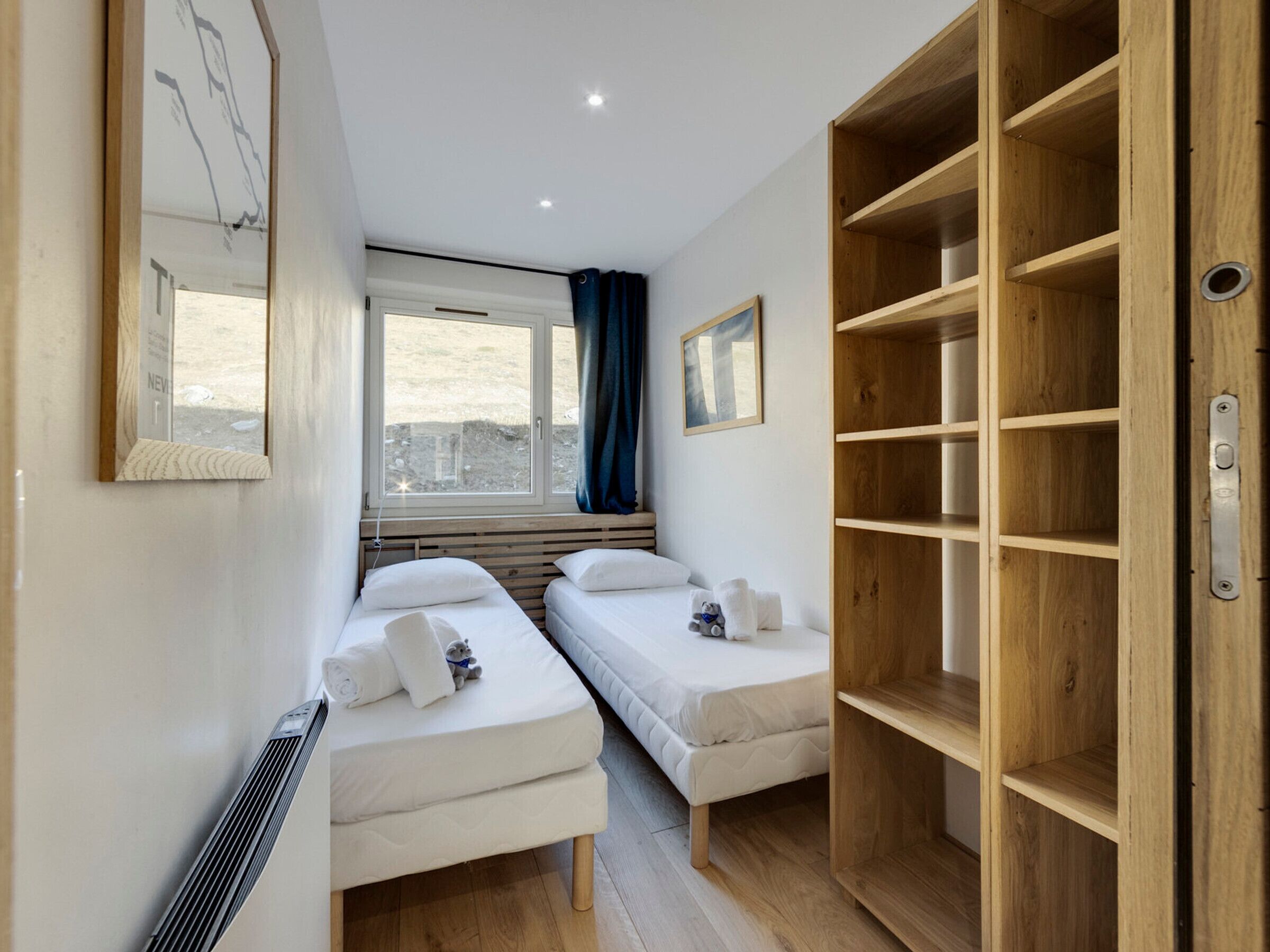 A small, modern room with two single beds, a large wooden wardrobe, a window with a view, and a ceiling light.