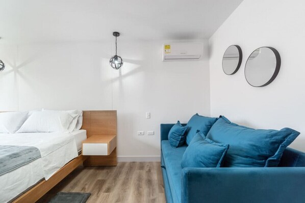 Une chambre moderne avec un lit, une table de chevet en bois, un canapé bleu, un appareil de climatisation et deux miroirs fixés au mur.