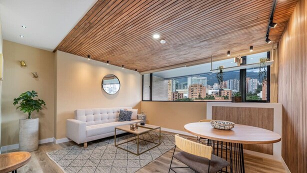 Una sala de estar moderna con un sofá blanco, una mesa de centro redonda y un comedor con una mesa y sillas redondas. Un ventanal ofrece vistas de la ciudad.