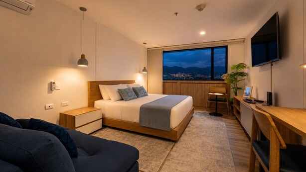 Ein modernes Hotelzimmer mit Bett, Nachttischen, einem Schreibtisch, einem Sessel, einem Fernseher und einem großen Fenster mit Blick auf die Stadt.