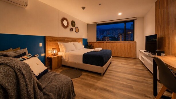 A modern bedroom with a city view, a bed, a desk, and a TV.