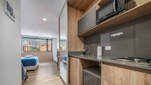 A compact kitchen with a microwave, sink, and stove. A bed and a window with a city view are visible in the adjacent room.