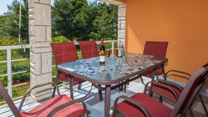 A covered outdoor patio with a glass table surrounded by red chairs, a bottle of wine, and a glass of wine on the table. - Apartments Horvatic (Vir)