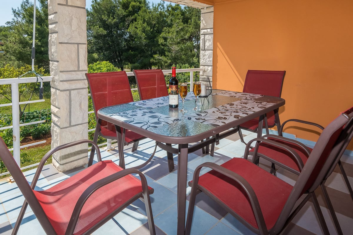 A covered outdoor patio with a glass table surrounded by red chairs, a bottle of wine, and a glass of wine on the table.