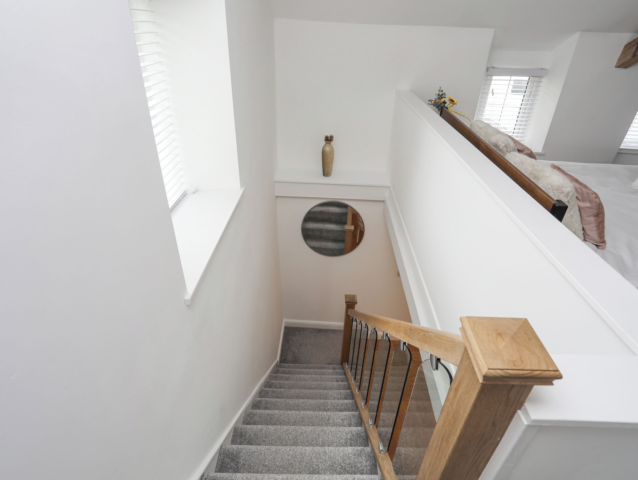 A staircase with a wooden handrail, carpeted steps, and a white wall with a small round mirror and a vase.