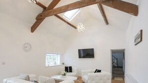 Ein geräumiges Wohnzimmer mit hohen Decken, Holzbalken, einem Flachbildfernseher, einer Uhr und einer Treppe, die zu einer weiteren Ebene führt.