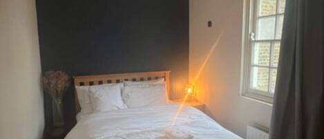 A neatly made bed with white linens and towels, a bedside lamp, a window with curtains, and a dark wall.