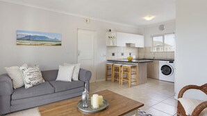 Ein kompakter Wohnbereich mit grauem Sofa, Couchtisch mit Kerzen, Küchenzeile mit Waschmaschine und Esstisch.