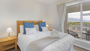 Ein ordentlich bezogenes Bett mit weißer Bettwäsche und Kissen in Blau-Weiß, eine Nachttischlampe und Blick auf einen Balkon mit Tisch und Stühlen.
