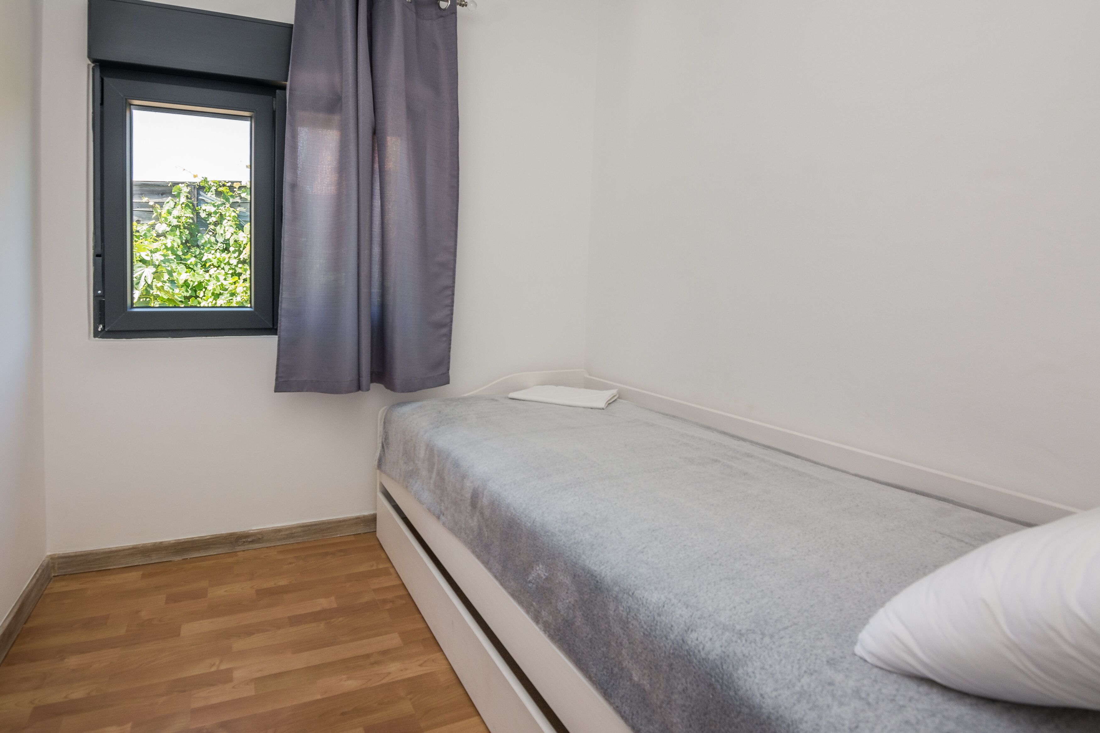 Una habitación pequeña con cama individual, ventana, suelo de madera y colcha gris.