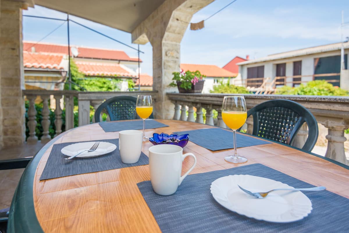 A round wooden table with a glass top, set for breakfast with two white mugs, a glass of orange juice, and a plate with cutlery.