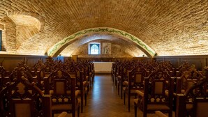 A church interior with wooden pews arranged in rows, a brick ceiling, and a central aisle leading to a stained glass window.