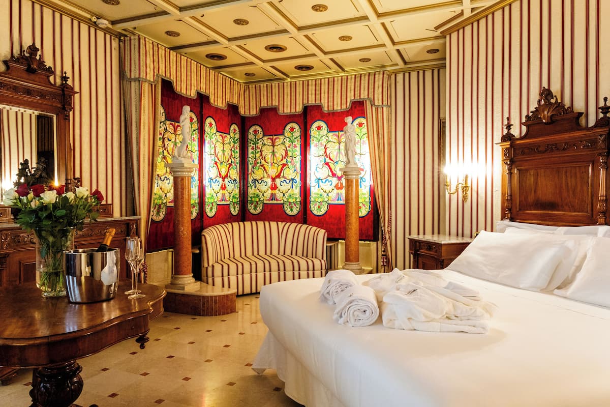 Ein Hotelzimmer mit einem großen Bett, einem Holz-Kopfteil, einer Sitzecke mit einem gestreiften Sofa und einem dekorativen Buntglasfenster.