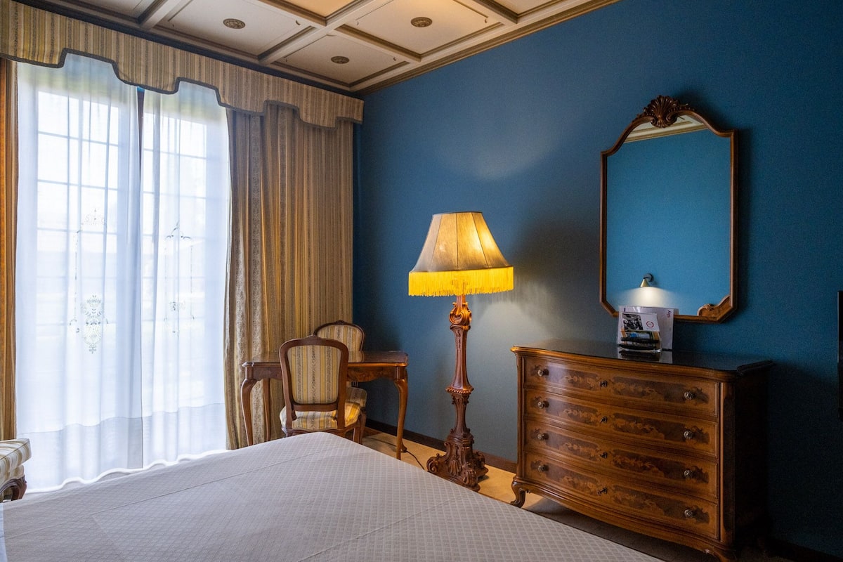 Ein Hotelzimmer mit einem Bett, einer hölzernen Kommode, einer Lampe, einem Sessel und einem großen Spiegel an der Wand.