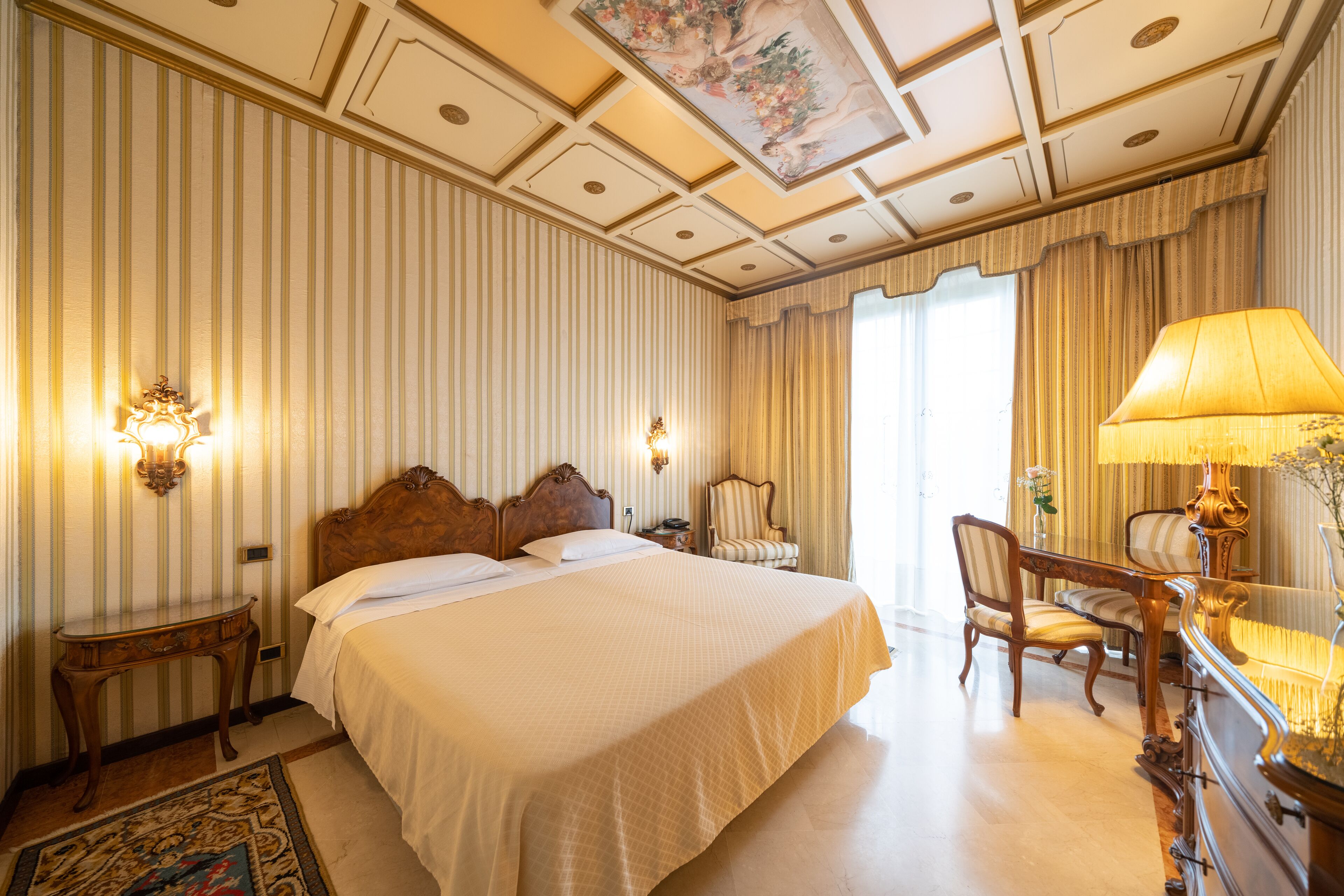A large bed with a wooden headboard, a nightstand, a desk with a lamp, and a chair.